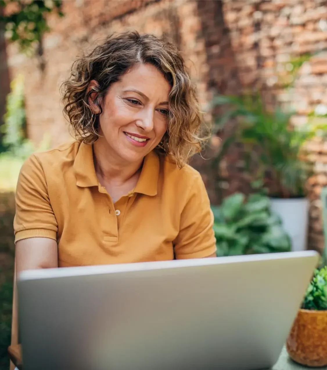 A relaxed woman is working on her laptop amidst a garden setting, sitting at a wooden table, embodying a balance of productivity and serenity in an outdoor space.