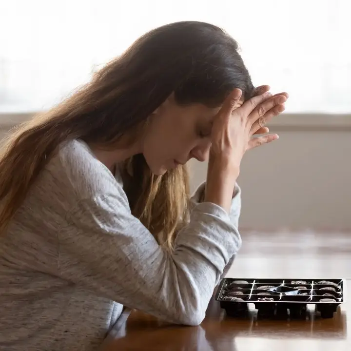 A woman sitting at a table with a box of chocolates, her hands resting on her head, looking stressed