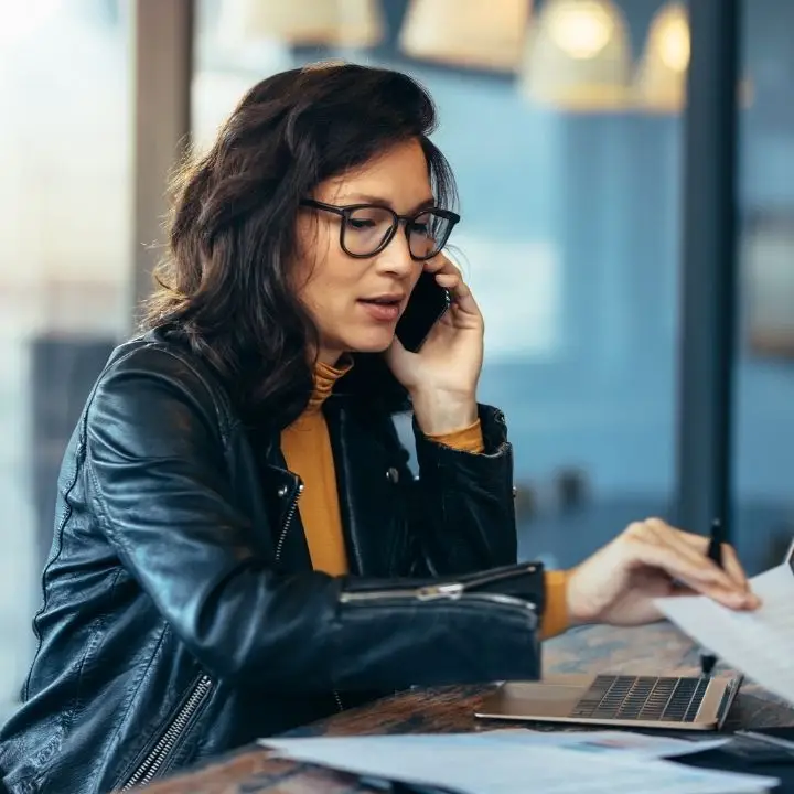 A woman talking on the phone while looking at papers on a table with a laptop