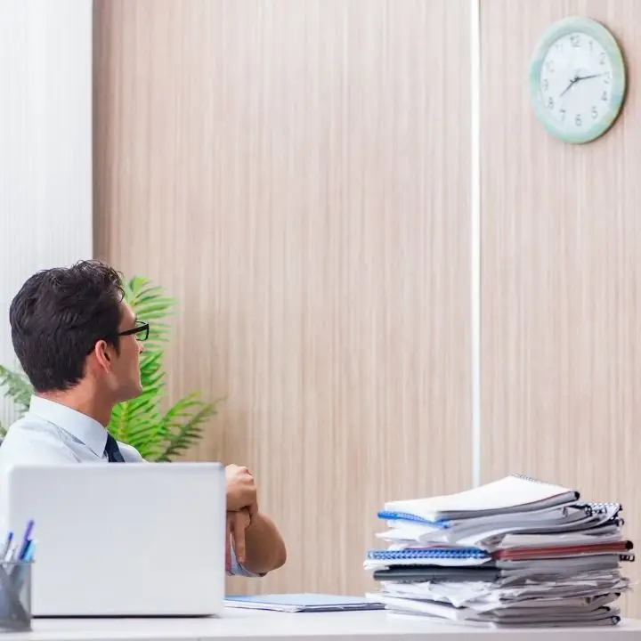 Man sitting at a desk looking up at a wall clock, with a stack of papers and a laptop in front of him.