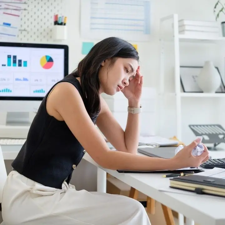 A woman at her workstation holding her forehead with one hand, looking overwhelmed.
