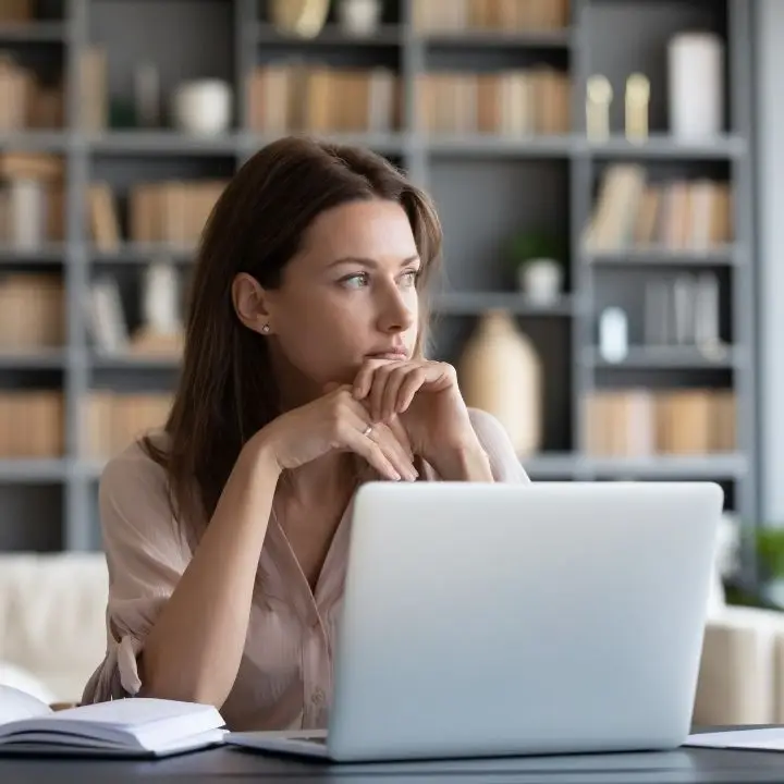 Adult woman sitting at laptop, looking away thoughtfully in a quiet workspace.