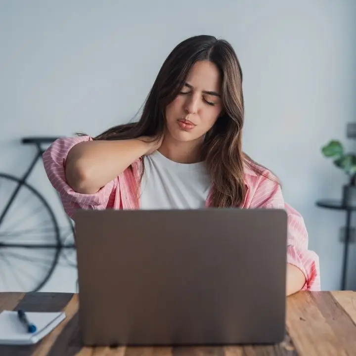 Woman working on laptop while rubbing her neck, showing muscle tension linked to anxiety.