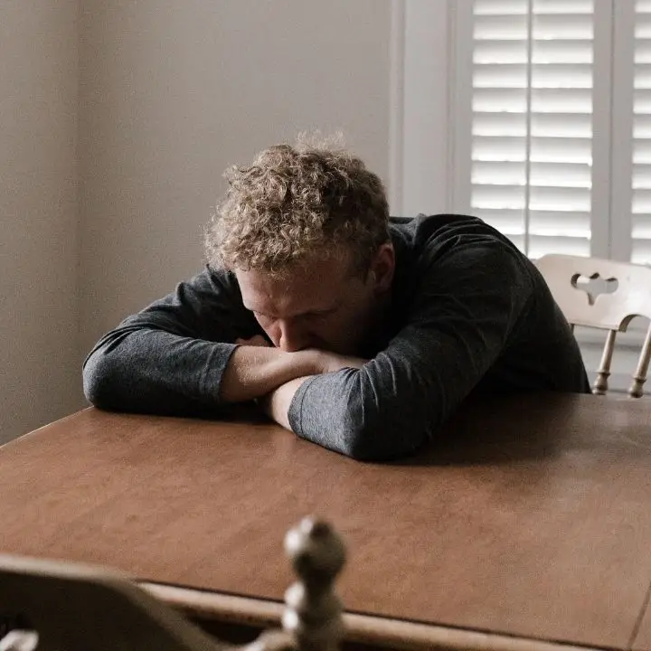 Man resting his head on folded arms at a table, appearing emotionally exhausted and overwhelmed by anxiety.