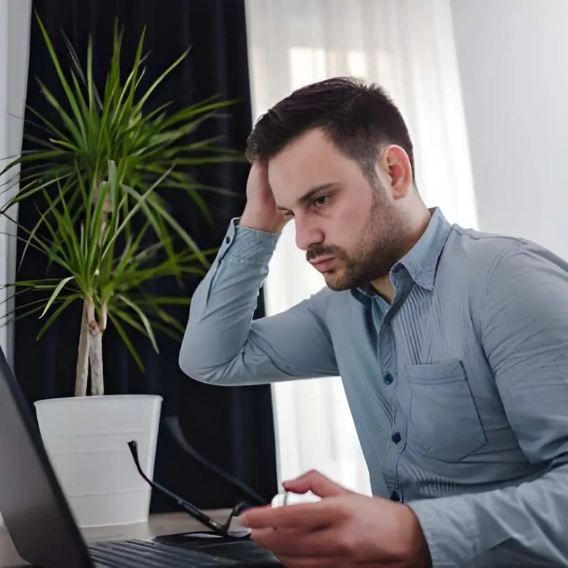 Man appearing stressed while working on a laptop beside a houseplant in an office.