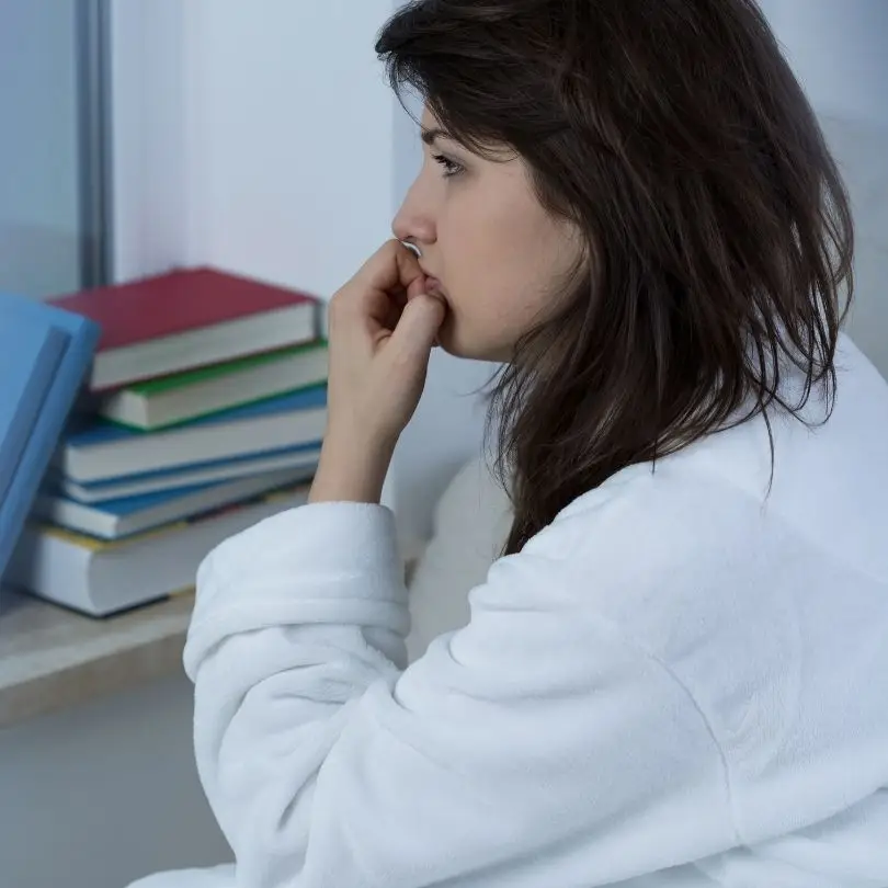 Woman sitting by a window in a robe, appearing worried and lost in thought.