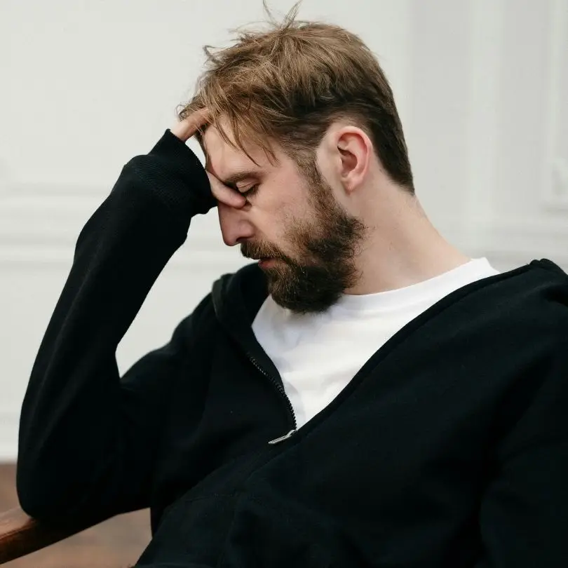 Man in black hoodie sitting with head in hand, eyes closed, appearing overwhelmed by obsessive thoughts.