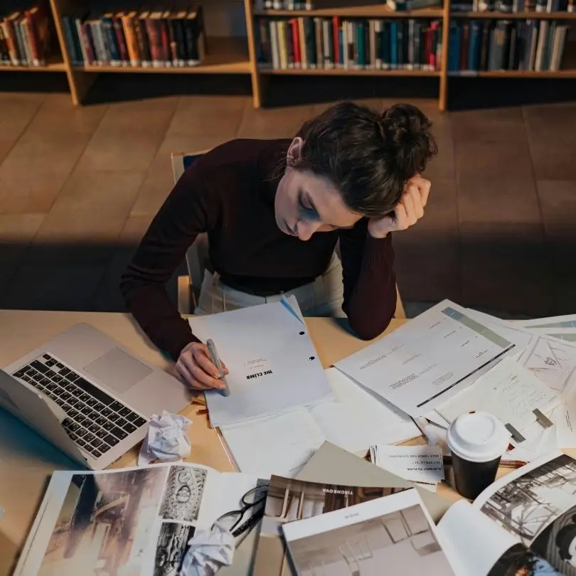 Perfectionist woman sitting at a desk surrounded by papers and a laptop, reviewing documents.